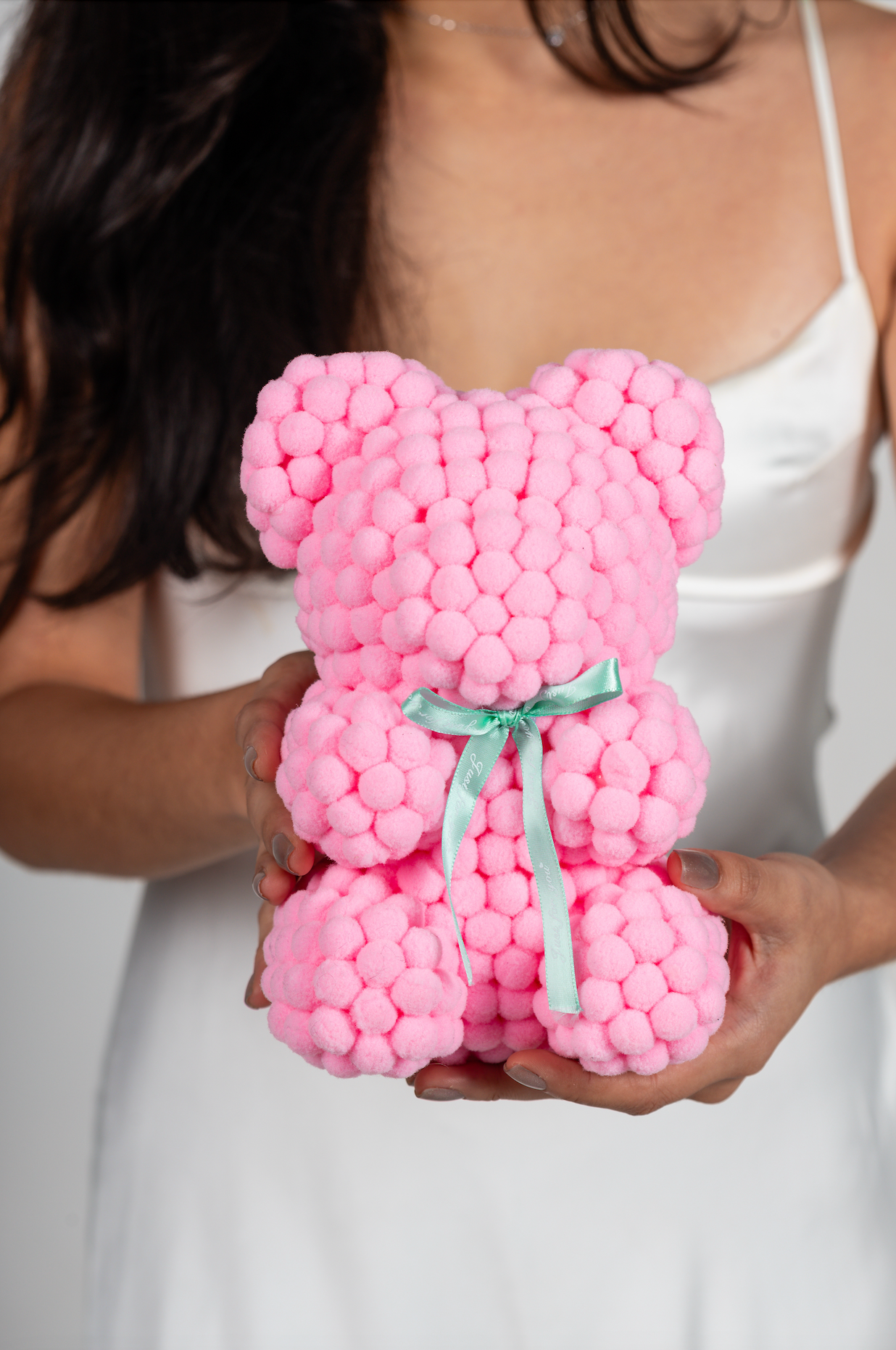 Bear made out of light pink-colored faux marshmallows. The bear is adorned with a green ribbon tied into a bow around its neck, and it is packaged in a clear-front gift box with a pink and white striped design. The top of the box features a teal and pink label that reads "MARSHMALLOW BEAR." This delightful creation is an excellent gift idea for various occasions such as anniversaries, birthdays, graduations, Christmas, Valentine's Day, or simply as a unique gift for someone special.